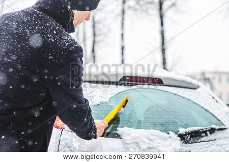 Young Man In Black Coat Cleans His Car With Yellow Brush During Snowfall. Winter Inclement Weather.