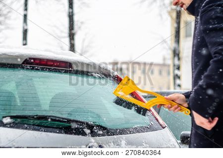 Young Man In Black Coat Cleans His Car With Yellow Brush During Snowfall. Winter Inclement Weather.