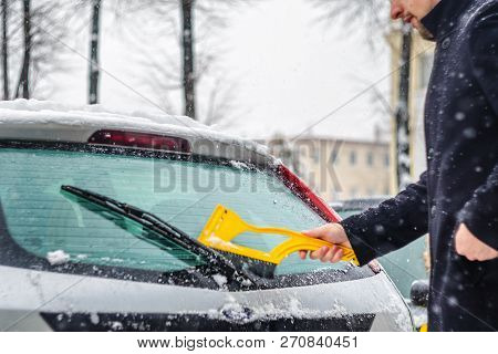 Young Man In Black Coat Cleans His Car With Yellow Brush During Snowfall. Winter Inclement Weather.