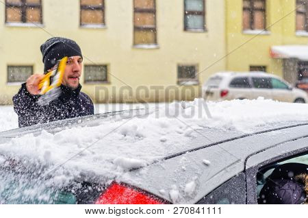 Young Man In Black Coat Cleans His Car With Yellow Brush During Snowfall. Winter Inclement Weather.