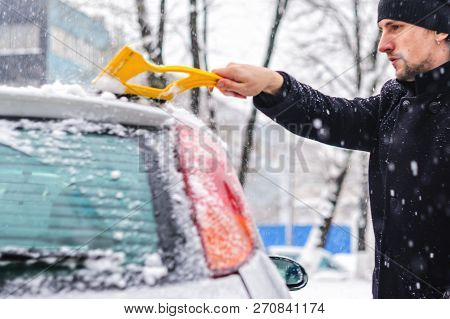 Young Man In Black Coat Cleans His Car With Yellow Brush During Snowfall. Winter Inclement Weather.