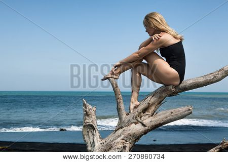 Girl With Slim Body In Swimsuit Sit And Relax On Old Tree Snag, Posing On Black Sand Sea Beach. Youn
