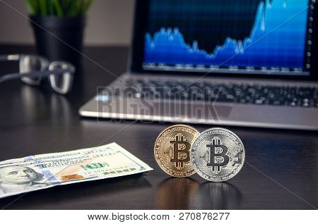 Bitcoins On The Table On The Background Of The Laptop With The Schedule, Next To The Dollars