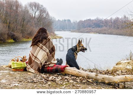 A Woman Covered With Warm Plaid Is Sitting Backside At Tha Bank River With A Dog By Her Side. The Co