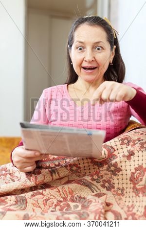 Positive Surprised Mature Woman Looks Newspaper At Home