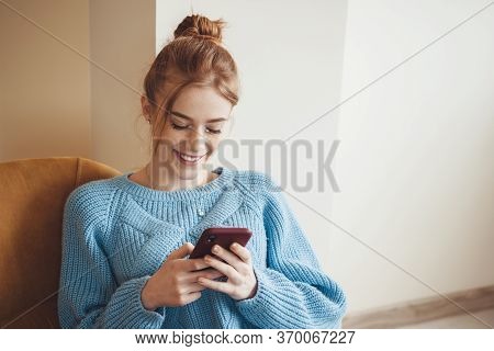 Ginger Lady With Freckles Is Smiling While Chatting On Mobile Wearing Blue Knitted Sweater At Home