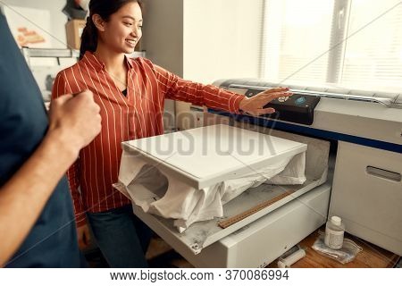 Young Workers, Man And Woman Are Preparing T-shirt For Printing In The Silk Screen Printing Machine 