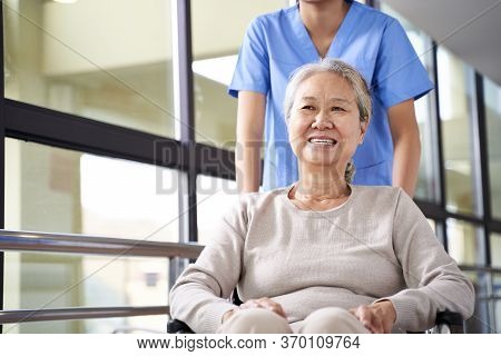 Asian Caregiver Assisting Wheelchair Bound Senior Woman In Hallway Of Nursing Home