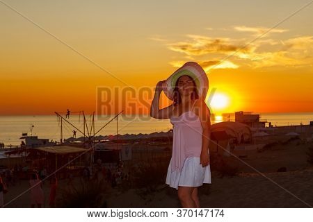 Woman In A White Dress And A Straw Hat On A Background Of The Sea At Sunset.