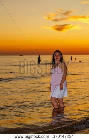 A Young Woman In A White Dress Stands On The Seashore At Sunset.