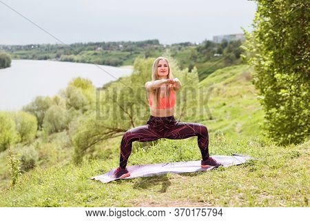 Fitness Instructor Is Doing Plie Squats On The River Bank Outdoors. Summertime.