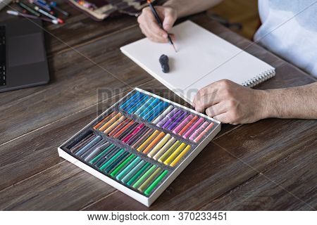 Person Painting With A Pastel Crayon Chalk On A White Paper With Pallette Of Pastel Chalks On The Wo
