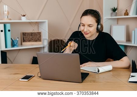 Smiling Happy Businesswoman In Headphones Sits At Desk, Looks At Laptop Screen, Making Notes, Partic