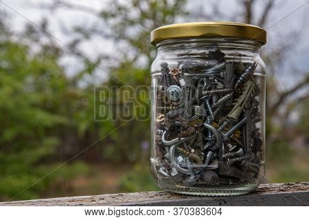 Transparent Glass Jar Of Assorted Hardware Bolts And Nuts Closeup Through Blurry Glass On Bokeh Back