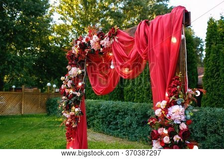 Wedding. Ceremony. Wedding Arch. Red Wedding Arch Of Flowers And Greenery Stands On The Green Grass 