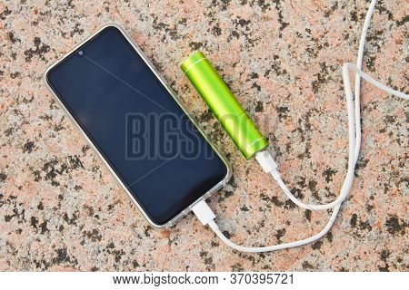 Smartphone And External Power Bank Lie On The Granite Surface In The Park While Charging