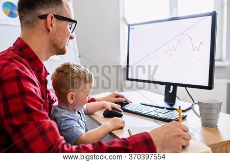 Father Works From Home Office While Babysitting With Toddler Kid. Dad Sits At The Desk With A Little