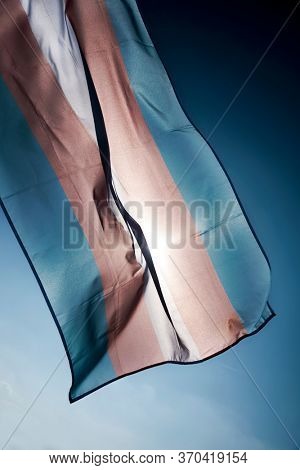 closeup of a transgender pride flag waving on the blue sky, moved by the wind, with the sun in the background