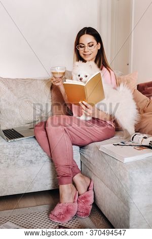 A Girl In Pajamas At Home Reads A Book With Her Dog Spitzer, The Dog And Its Owner Are Resting On Th