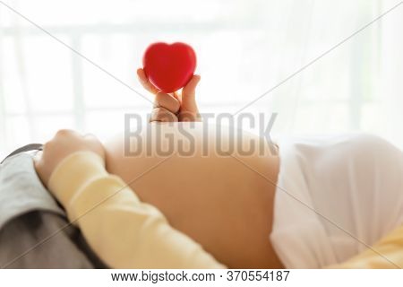 Happy Pregnant Young Woman Holding Red Heart In Hand On Top Of Mother Belly In Hospital Bed. Prenata