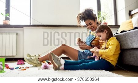 African American Woman Baby Sitter Entertaining Caucasian Cute Little Girl. They Are Playing With Do