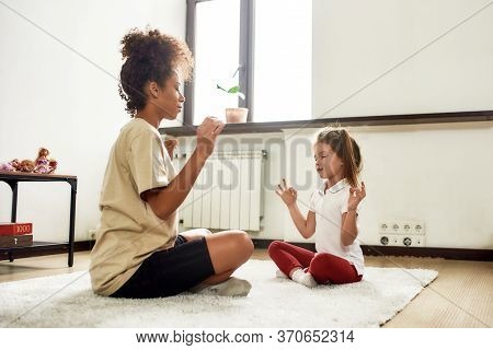 Full Length Shot Of African American Woman Baby Sitter And Caucasian Cute Little Girl Doing Yoga At 