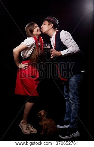 Funny Oktoberfest Couple In National Ethnic Dress With Mugs Of Beer, Small Shaggy Dog Kisssing On Bl