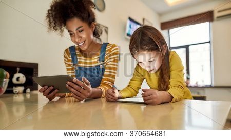 African American Woman Baby Sitter And Caucasian Cute Little Girl Reading, Using Tablet Pc, Sitting 