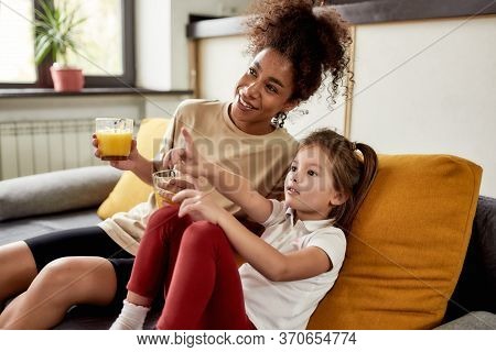African American Woman Baby Sitter Entertaining Caucasian Cute Little Girl. They Are Sitting On The 