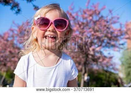 Cute Little Baby Girl Laugthing In Pink Glasses At Summer, Looking At Camera. Happy Blonde Caucasian