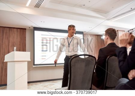 Asian Male Speaker In Suit Giving A Talk At Corporate Business Meeting, Forum. Chart On The Screen I