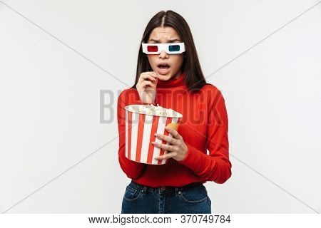Photo of uptight brunette woman wearing 3D glasses holding popcorn bucket isolated over white background