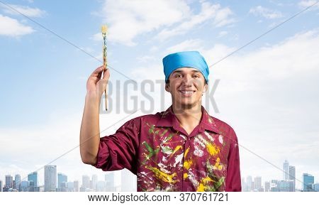 Smiling Young Painter Artist Pointing Upwards With Paintbrush In Hand. Portrait Of Happy Painter On 