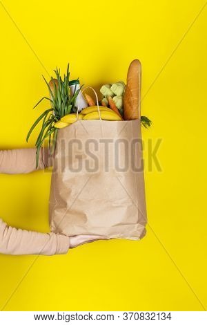 Food delivery or donation concept. Grocery store shopping. Girl or woman holds a paper bag filled with groceries such as fruits, vegetables, milk, yogurt, eggs isolated on yellow.