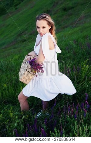 Beautiful Girl In White Dress With Straw Bag And Wildflowers In Meadow.