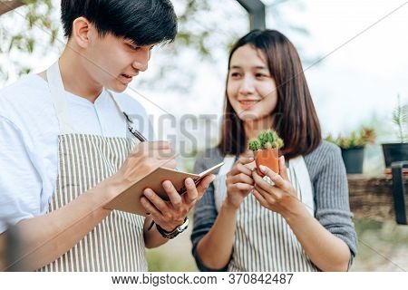 Women Hold Cactus And A Man Writing Note In A Book. Love Couple Enjoy Hobby With Darden Cactus.