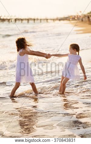 Girl Children In White Dresses Playing On The Beach. Older Sister Pulls The Younger Into The Water. 