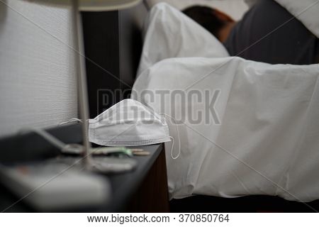Asian Male Sleeping In Bed Feeling Sick With Covid-19. Non-surgical Face Mask On Nightstand Is Cente