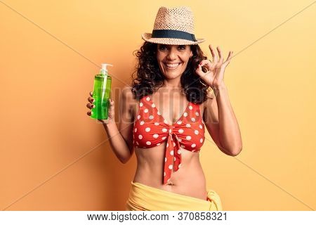 Middle age beautiful woman wearing bikini and hat holding aloe vera lotion doing ok sign with fingers, smiling friendly gesturing excellent symbol 