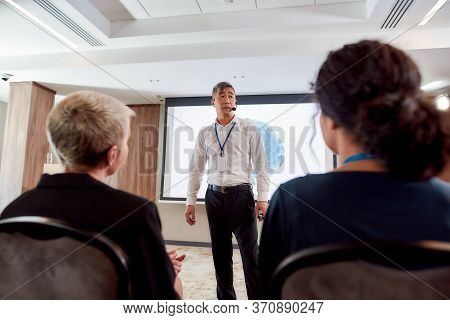 Asian Male Speaker In Suit Giving A Talk At Corporate Business Meeting, Forum. Audience Listening To
