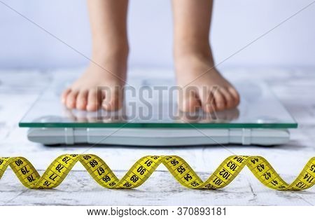 Weight Control Concept With Centimeter In Focus And Blurred Kid's Feet On Digital Scale In The Backg