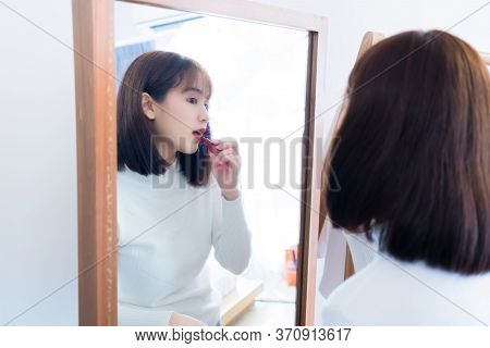Asian Woman Choosing Make Up With Cosmatic Applying Lipstick And Smiling Happy In Front Mirror At Ho