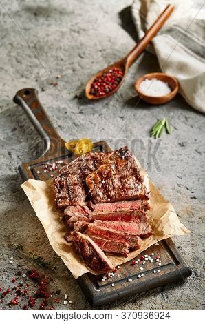 Classic ribeye steak served in waxed paper on wooden cutting board. Homemade barbecue dish concept. Roasted beef steak with yellow chilli pepper slices and salt crumbs. Rustic spoon and spice bowl