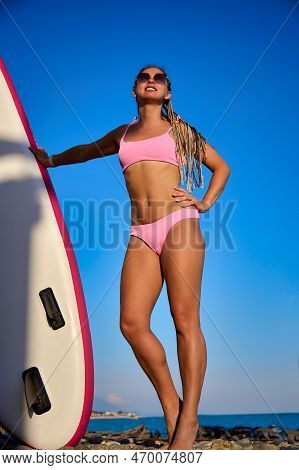Tanned Slender Girl In A Pink Swimsuit Stands Against A Blue Sky Near A Sapsurf Board. Cute Surfer P