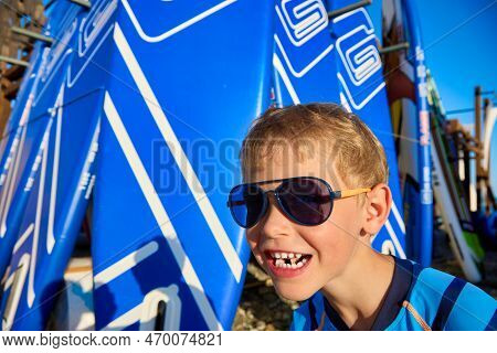 Close-up Of A Grimace Of A Little Boy In Black Sunglasses Against The Backdrop Of A Sap Board Rack O