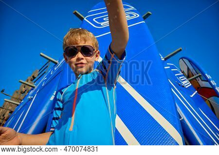 Caucasian Child Surfer Pours Sand On The Beach In Summer On The Background Of Swimming Boards. A Coo