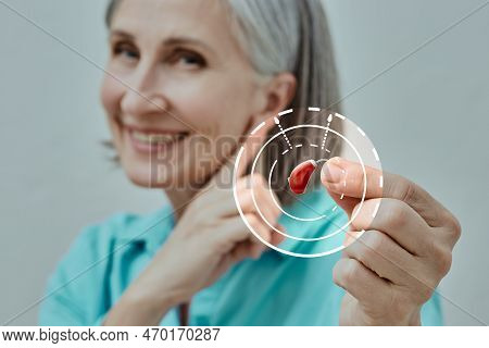 Mature Woman Holding Bte Hearing Aid In Hand On Foreground And Points To Her Ear, Concept. Hearing L