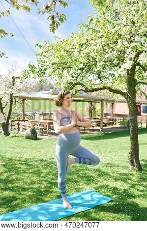 Outdoor Portrait Of Happy Young Pregnant Woman Practicing Youga In Spring Garden Under Blooming Appl