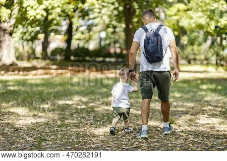 Family Walk In The Forest. A Grown Man Walks And Holds The Hand Of A Little Boy Who Has The Same Clo
