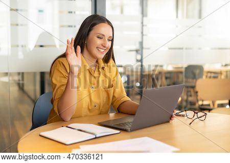 Video Meeting Concept. Cheerful Young Woman Has Video Call On The Laptop In Office, Waving Hi Into W